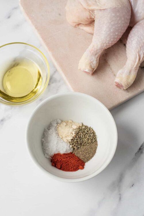 Seasoned raw whole chicken and spices on a marble countertop, ready for roasting or cooking.
