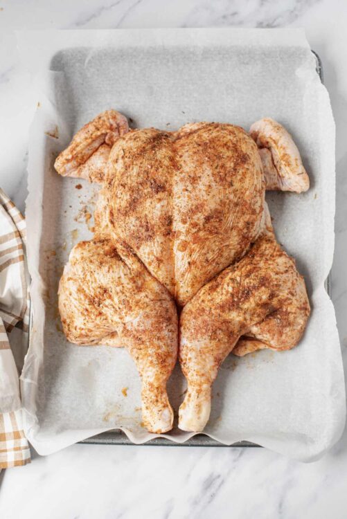 Seasoned whole raw chicken on baking sheet with parchment paper, ready for roasting.