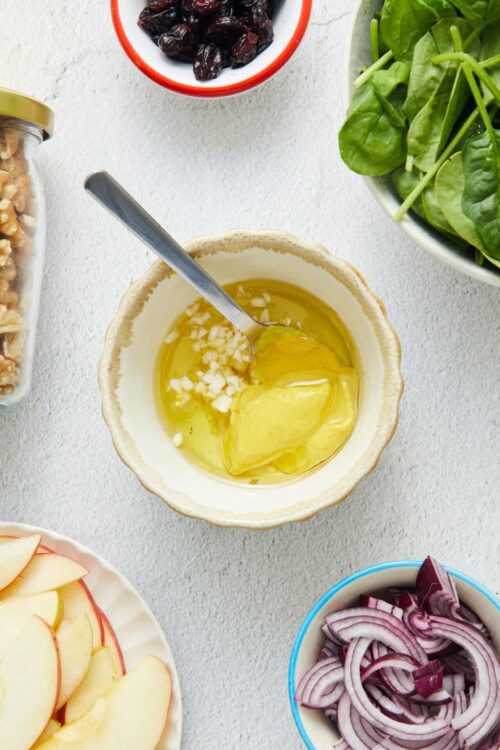 Fresh salad ingredients including chopped garlic, olive oil, spinach, red onions, dried cranberries, and sliced apples.