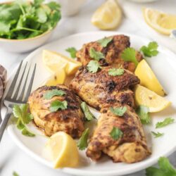 Grilled chicken thighs with lemon wedges and fresh cilantro on a white plate for a flavorful meal.