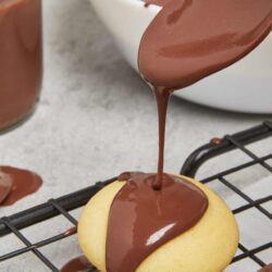 Creamy chocolate frosting being poured over a vanilla cookie on a cooling rack.