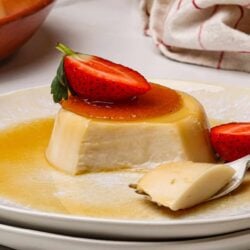 Creamy caramel flan with strawberries on a white plate, served with caramel sauce and a spoonful.