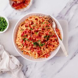 Creamy tomato bacon pasta topped with fresh herbs on a white bowl on marble surface.