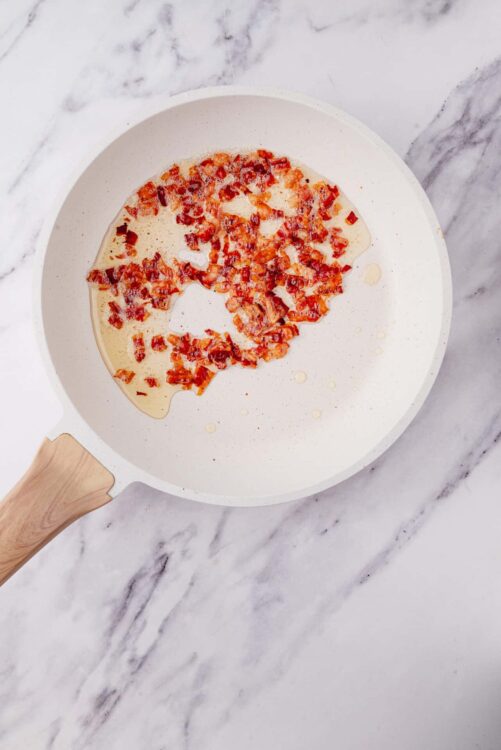 Savory bacon pieces in creamy sauce in white skillet on marble surface.