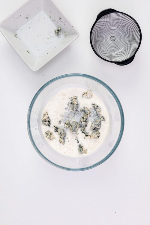 Crumbled blue cheese in a mixing bowl with heavy cream on white background.