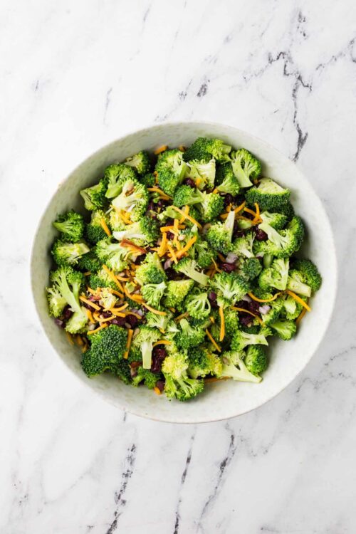 Fresh broccoli salad with shredded cheese and dried cranberries in a white ceramic bowl on marble surface.