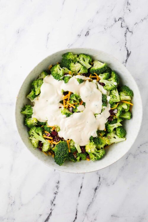 Crispy broccoli salad topped with creamy dressing and shredded cheese in a white bowl.