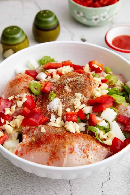 Seasoned chicken thighs with chopped vegetables in a white bowl for baking or roasting.