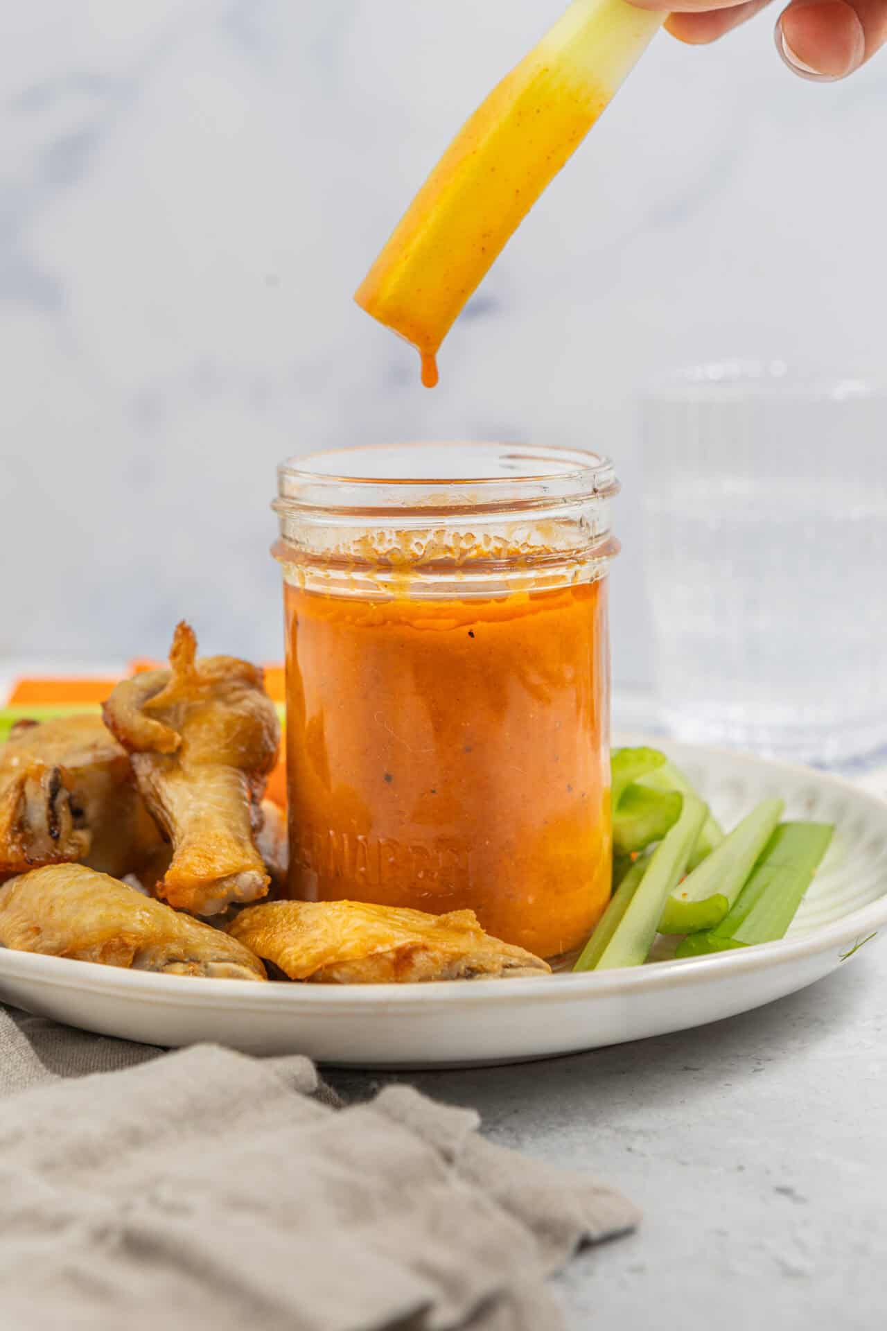 Golden mango dipping sauce being poured into a jar with chicken wings and celery on a white plate.