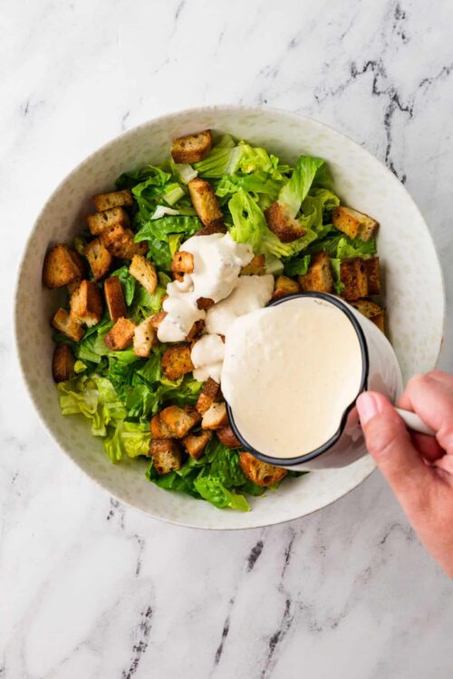 Creamy salad dressing being poured over a fresh chicken Caesar salad with romaine lettuce and croutons in a white bowl.