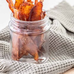 Crispy homemade bacon jerky in a glass jar on a kitchen towel. Perfect for snacks, made from perfectly cured and smoked bacon.
