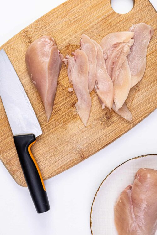 Sliced raw chicken breasts on a wooden cutting board with a chef's knife.