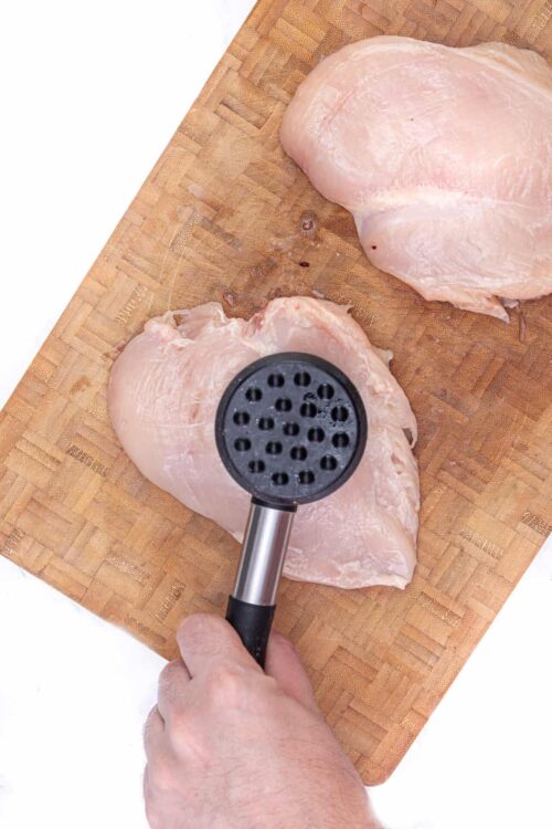 Raw chicken breasts on a wooden cutting board with a brief meat tenderizer.