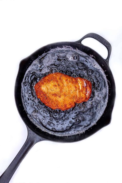 Crispy breaded chicken cutlet frying in cast iron skillet with bubbling oil on white background.