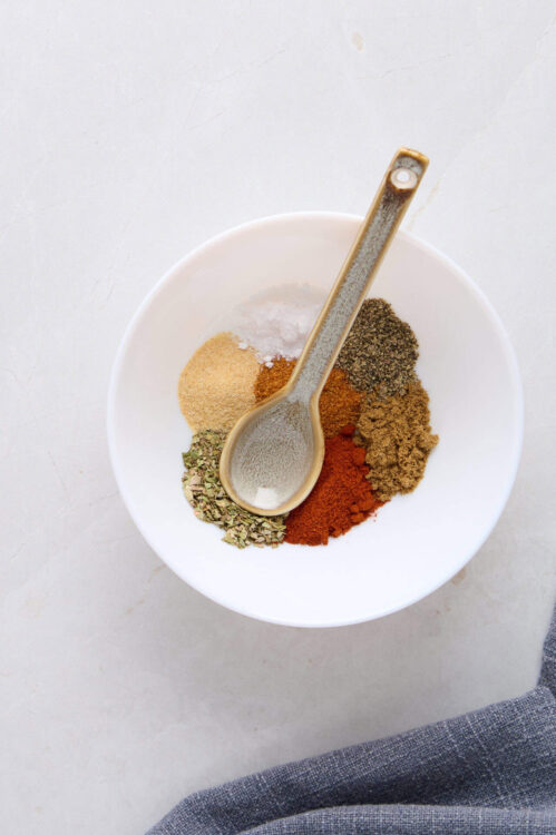 Ground spices and herbs in a white bowl with a spoon on a light background.