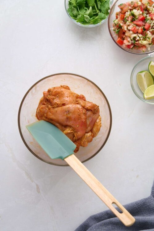 Marinated chicken in glass bowl with fresh ingredients around, ready for cooking.