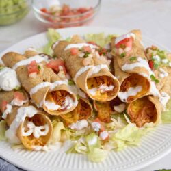 Crunchy beef and cheese taquitos served on a white plate garnished with shredded lettuce and topped with sour cream and salsa.