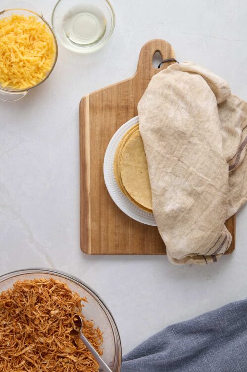 Flour tortillas on a white plate covered with a linen cloth, surrounded by shredded chicken, cheese, and cooking ingredients, ready for a homemade taco or burrito recipe.