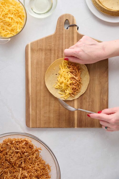 Shredded chicken and cheese being assembled in a taco on a wooden cutting board.