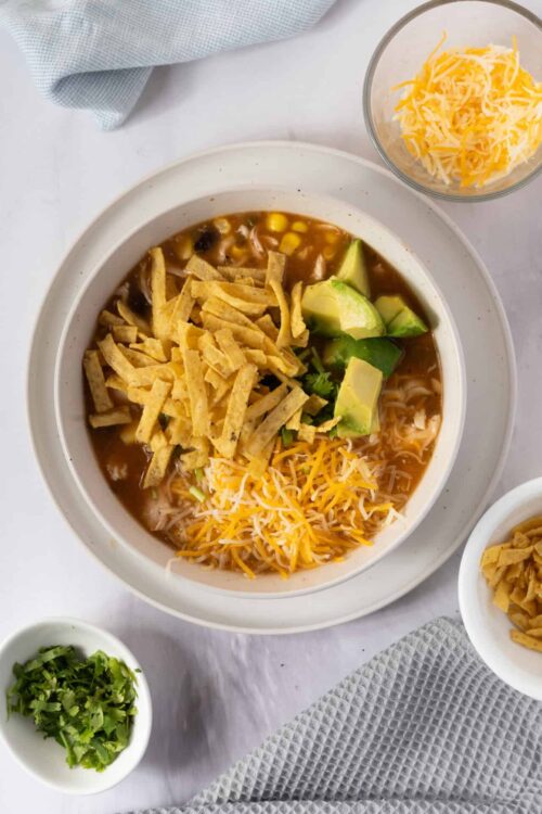Creamy chicken tortilla soup with shredded cheese, avocado, and crispy tortilla strips in a white bowl.