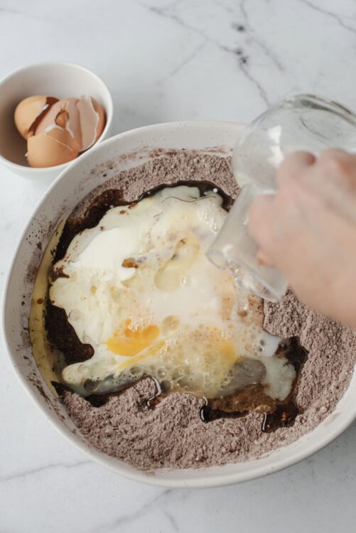 Eggs being added to a bowl of dry cake mix for baking, with cracked eggshells in a small dish nearby.