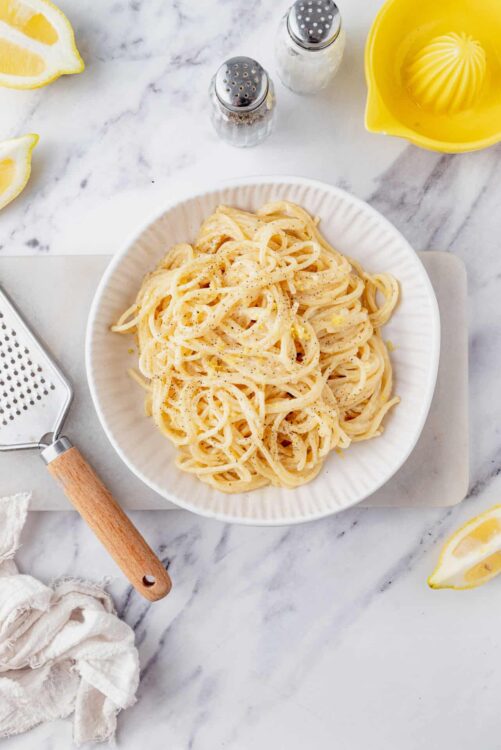 Creamy lemon pasta in a white bowl with lemon wedges and seasoning on a marble surface.