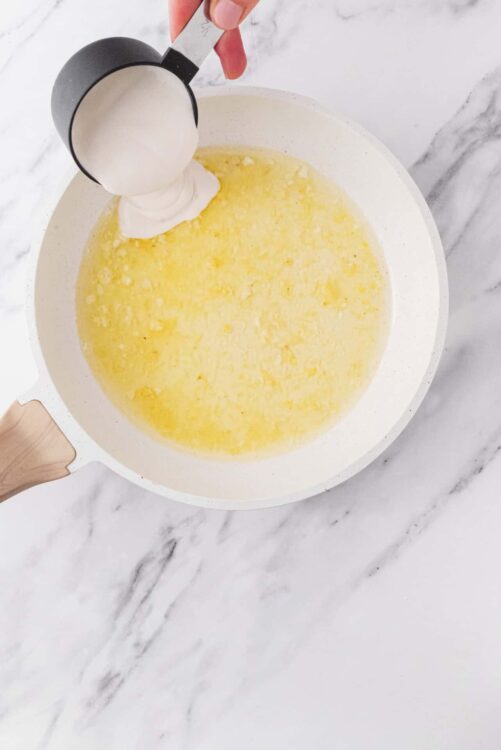 Cream being poured into a white skillet with melted butter on marble countertop.