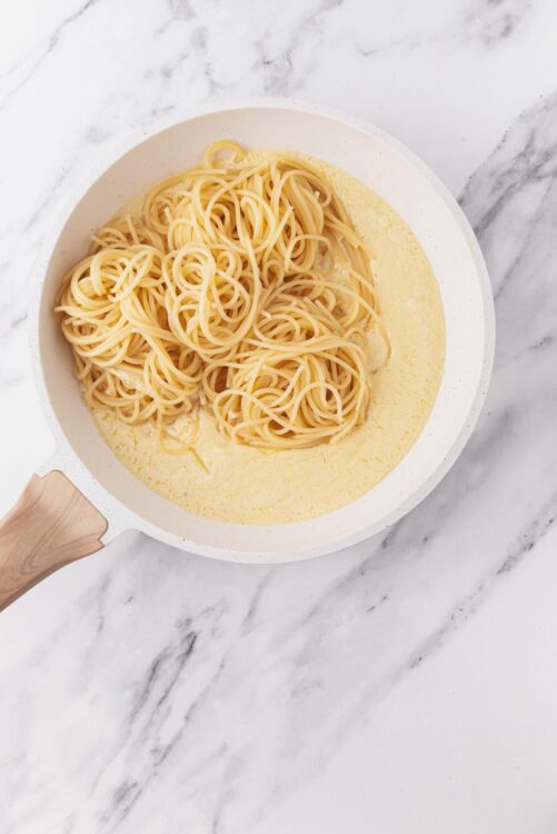 Creamy spaghetti in a white skillet with a wooden handle on marble surface.