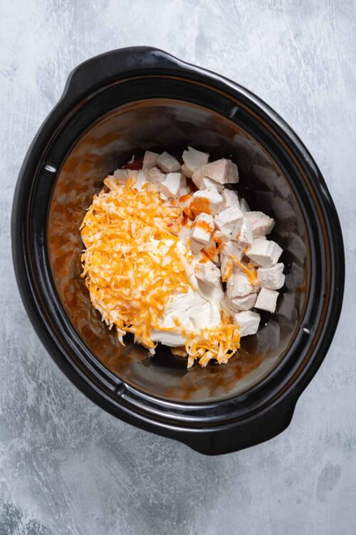 Shredded cheddar cheese and cooked chicken in a slow cooker for recipe preparation.