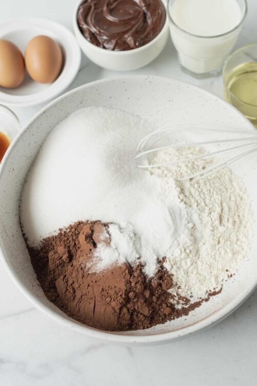 Flour, cocoa powder, and sugar for baking, with eggs, milk, and chocolate in the background.