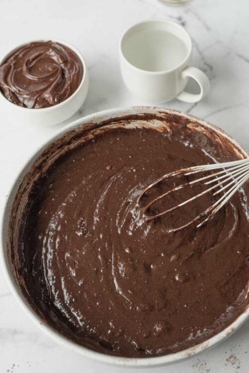 Fertile chocolate cake batter in large mixing bowl with whisk, on white marble surface.