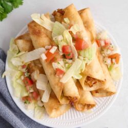 Crispy baked chili lime chicken taquitos with fresh salsa and lettuce on a white plate. Perfect for quick and flavorful appetizers or snacks.