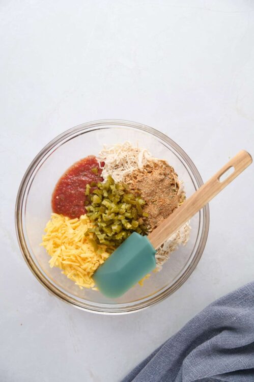 Cream cheese, shredded cheese, diced pickles, tomato sauce, and spices in a glass bowl for recipe prep.