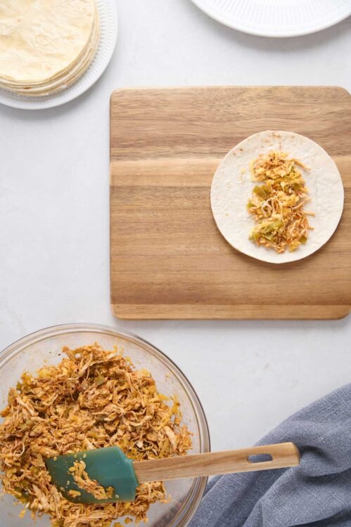 Shredded chicken filling on a wooden cutting board with tortillas and mixing bowl.