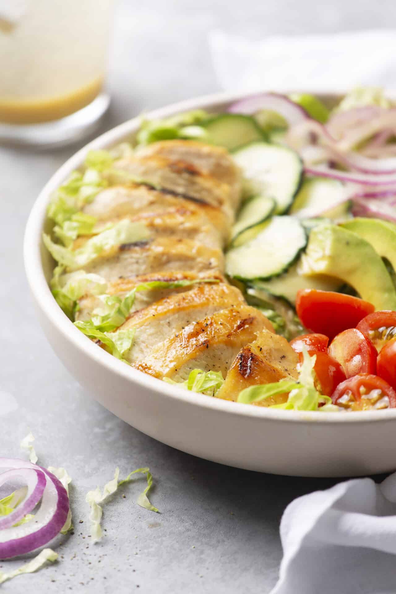 Fresh grilled chicken salad with cucumbers, cherry tomatoes, red onions, and crisp lettuce in a white bowl.