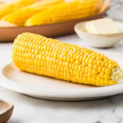 Fresh sweet corn on a white plate with butter and sliced corn in the background, perfect for summer recipes.