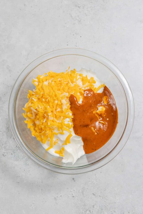 Cream cheese, shredded cheddar, and spicy buffalo sauce in a glass bowl for cheesy buffalo chicken dip.