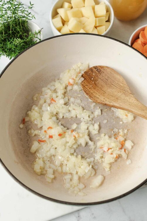Sautéing onions for a vegetable-based recipe in a white enamel cast iron Dutch oven. Fresh vegetables and herbs are visible around.
