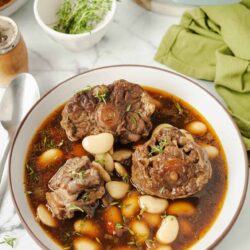 Succulent beef stew with white beans in a brown broth, garnished with herbs, on a white plate.