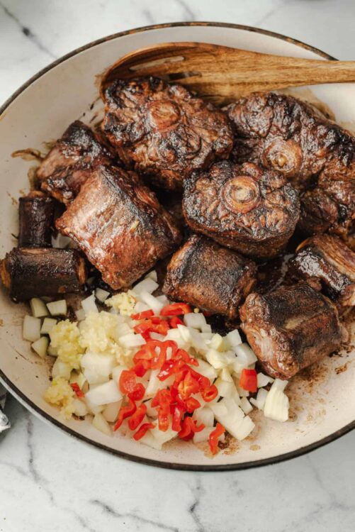 Braised beef short ribs with garlic, onions, and red chilies in a skillet.