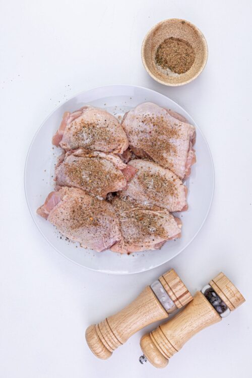 Seasoned raw chicken thighs on a white plate with salt and pepper; baking ingredients for delicious homemade recipes.
