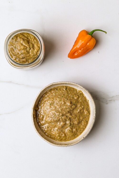 Spicy green salsa in a small bowl next to a jar of salsa and a yellow chili pepper on white background, healthy condiment.