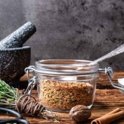 Seasoning spice blend in glass jar on rustic wooden surface with mortar and pestle, cinnamon stick, and fresh herbs.