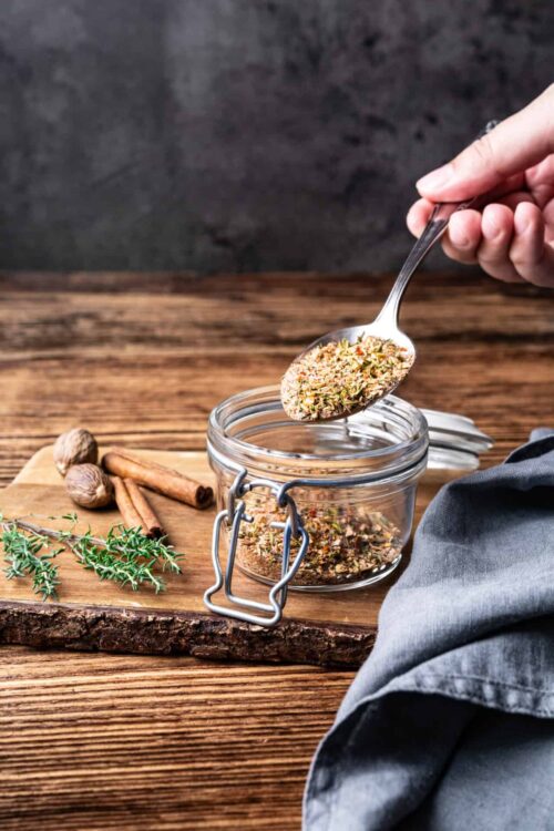 Dried herb mixture being scooped into a glass jar with cinnamon sticks and thyme on a rustic wooden surface.