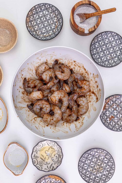 Succulent cooked shrimp in a marinade in a white bowl, surrounded by decorative bowls and salt jar.