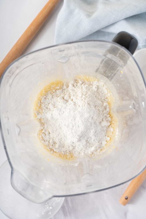 Flour being added to a mixing bowl with cake batter, on a light-colored surface.