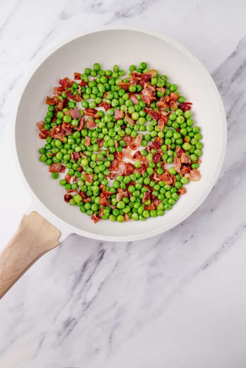 Fresh green peas and crispy bacon cooking in a white skillet, perfect for flavorful side dishes or breakfast recipes.