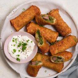 Crispy egg roll slices filled with vegetables and meat served with dipping sauce, garnished with green onions.