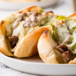 Buttery bread rolls filled with steak, peppers, onions, and melted cheese on a white plate.