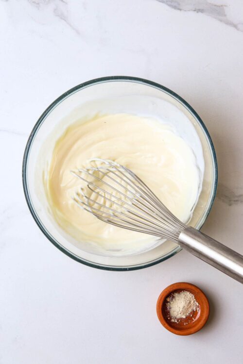Creamy homemade mayonnaise in a glass bowl with a whisk, on a marble surface.